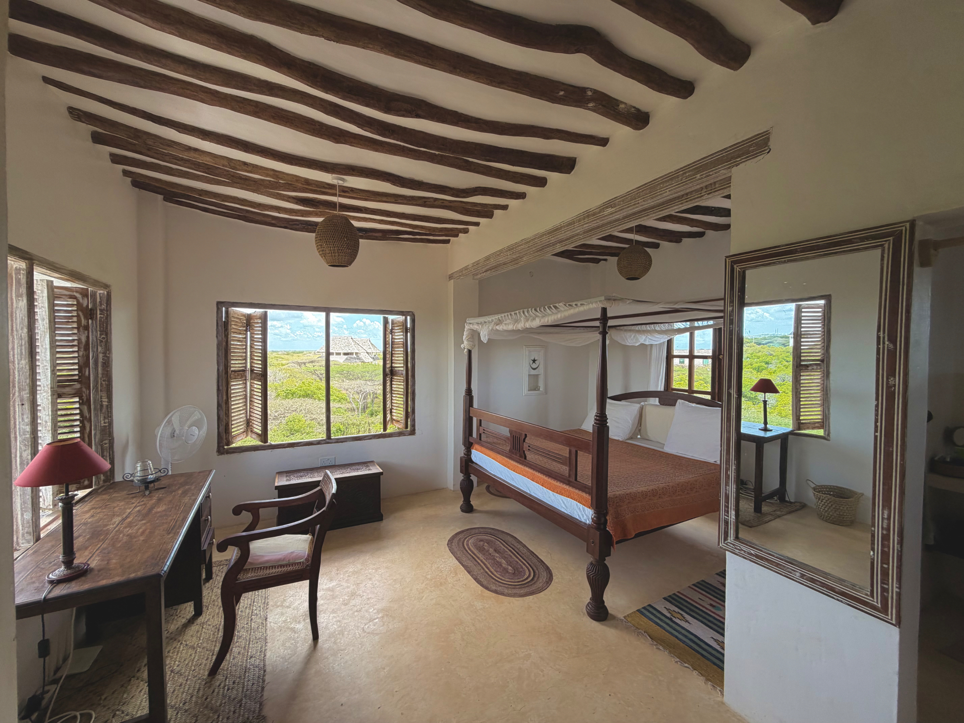 A bright bedroom at Mkoko House with a four-poster bed, wooden desk, shuttered windows and mangrove-pole beams