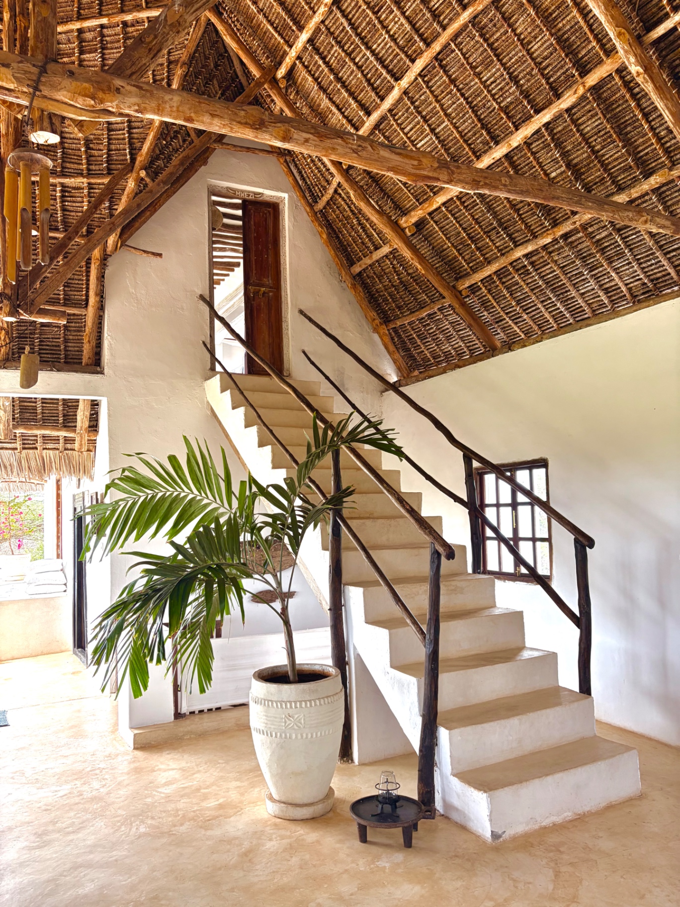 Whitewashed staircase with mangrove-pole railings beneath a high makuti roof, with a potted palm in the hall