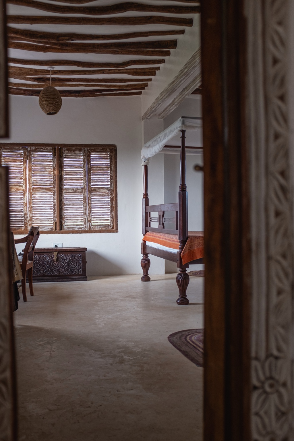 A bedroom at Mkoko House glimpsed through a carved timber doorway, with louvred shutters beyond