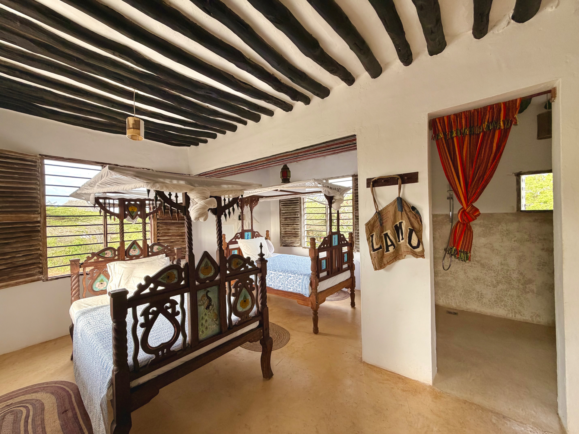 The Mkoko room with two carved four-poster beds under dark mangrove-pole beams, an open shower beyond a striped curtain